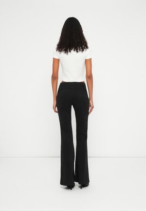Femme aux cheveux bouclés, debout face à un mur blanc, portant un haut blanc à manches courtes et un pantalon noir évasé avec des talons noirs.