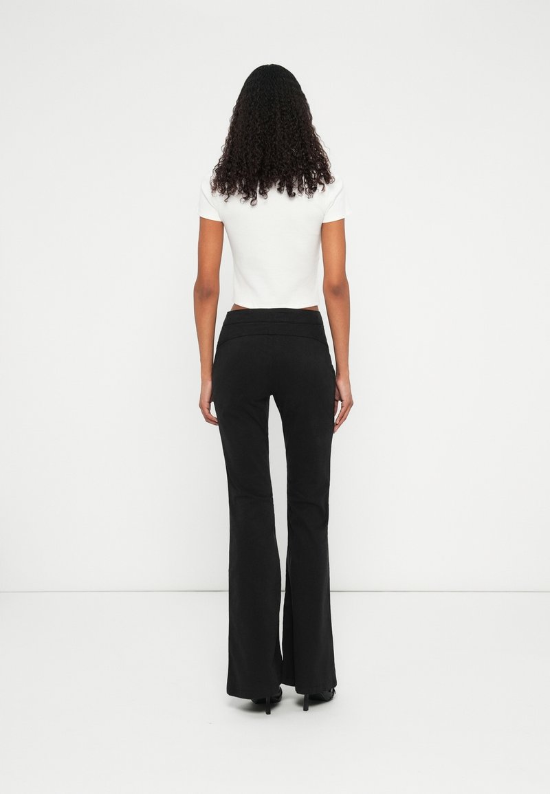 Femme aux cheveux bouclés, debout face à un mur blanc, portant un haut blanc à manches courtes et un pantalon noir évasé avec des talons noirs.