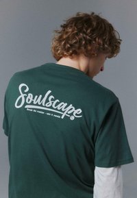 T-shirt en coton vert à coupe décontractée, avec un texte blanc au dos : "Soulscape" et "Brisez la routine - sortez dehors." Manches courtes.