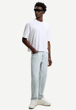 Tricou alb din bumbac cu umeri lăsați, blugi din denim de culoare albastră deschisă și adidași albi. Modelul poartă ochelari de soare negri și un colier din argint.