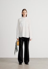 Fehér hosszú ujjú gombos ing, fekete széles szárú nadrág, kis sárga táska és mintás sál tartva. Minimalista dizájn.