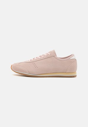 Zapatilla deportiva de ante rosa claro con punta redondeada, parte superior texturizada, acento blanco en el talón y suela de goma color caramelo. Presenta perforaciones sutiles para mayor transpirabilidad.