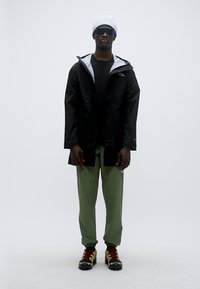 Μαύρο αδιάβροχο μπουφάν με γκρι φόδρα, πράσινο παντελόνι και μαύρα με κόκκινα παπούτσια. Αξεσουάρ περιλαμβάνουν ένα λευκό σκούφο και ανακλαστικά γυαλιά ηλίου.