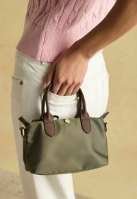 Malá olivovo zelená kabelka s hnedými koženými detailmi a dvojitými rúčkami, držená v ruke. Nosí sa s ružovým pleteným topom a bielymi nohavicami.