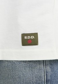 White cotton t-shirt featuring a green patch with "R.D.D." embroidered in white and a red circle. Cotton fabric, short sleeves, simple design.