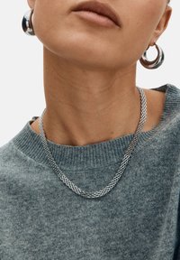 Silverkedja med sammanlänkade länkar, bärs över en grå stickad tröja med rundad halsringning och synlig struktur. Stor silverring i örat.