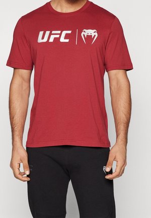 Hombre vistiendo una camiseta roja de UFC con logo blanco y pantalones cortos negros, de pie contra un fondo claro y liso.
