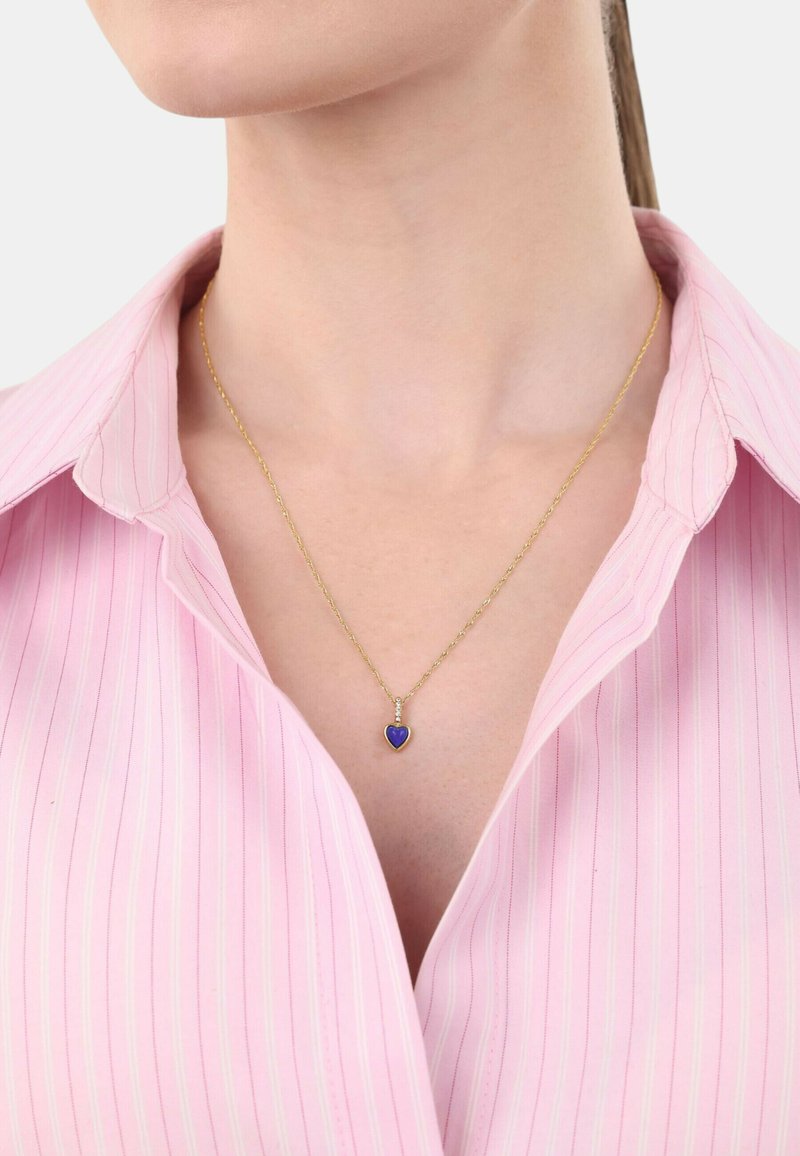 Collar de cadena de oro con un colgante en forma de corazón púrpura, llevado sobre una camisa a rayas rosas. El colgante tiene un acabado suave.