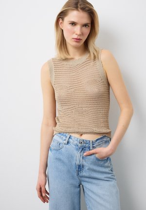 Jonge vrouw met blond haar, gekleed in een beige mouwloos gebreid topje en lichtblauwe, hooggetailleerde spijkerbroek, staand met één hand in haar zak.