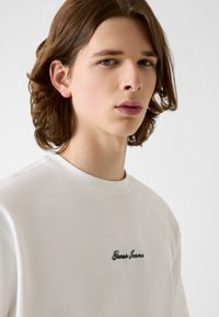 Biała teksturowana bluza z czarnym haftem "Guess Jeans" na piersi. Posiada okrągły dekolt oraz miękki, gładki materiał.