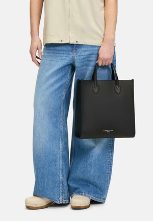 Schwarze Ledertasche mit strukturiertem Design, doppelten Griffen und dezentem Logo. Kombiniert mit einem hellbeigen Hemd und weit geschnittenen blauen Jeans.