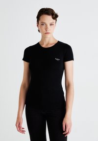 Tânără cu păr scurt, creț, purtând un tricou negru ajustat și pantaloni negri, stând în fața unui fundal alb simplu.