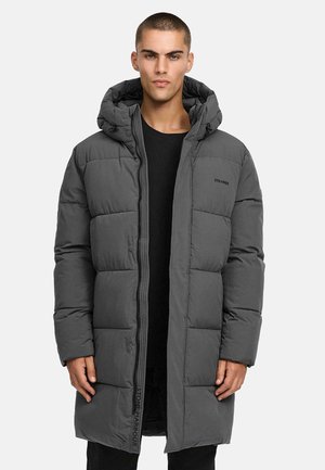 Man in een lange, donkergrijze gewatteerde winterjas met een capuchon, opengehouden over een zwart shirt, staand tegen een effen lichte achtergrond.