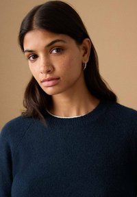 Pulover albastru închis cu decolteu rotund, manșete striate și textură moale. Modelul are părul lung și închis la culoare și poartă cercei mici argintii tip cerc.