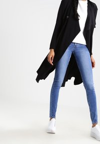Black, long cardigan with an asymmetrical hem, layered design, and single button detail. Paired with blue skinny jeans and white sneakers.