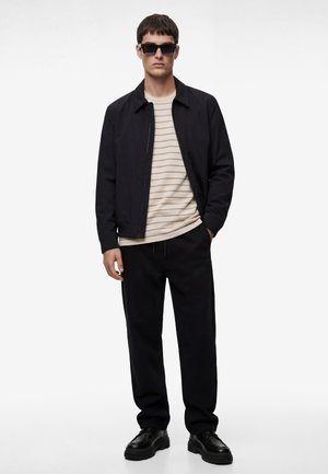 Mann trägt schwarze Jacke, schwarze Hose, schwarze Schuhe, gestreiftes beiges Hemd und schwarze Sonnenbrille, steht mit einer Hand in der Hosentasche.