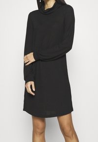 Schwarzes Kleid aus weichem Stoff, mit einem Wasserfallausschnitt, langen Ärmeln und einer lockeren Passform, das bis zur Mitte des Oberschenkels reicht. Einfaches Design, ohne Muster.