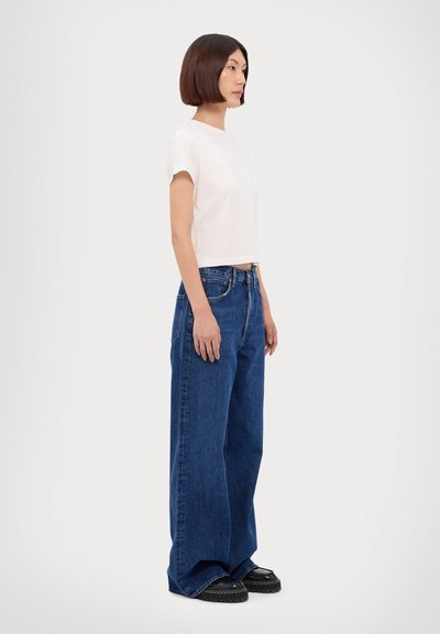 Persona joven con el pelo corto, que lleva una camisa blanca de manga corta, vaqueros azules de pierna ancha y zapatos negros gruesos, de pie de lado.