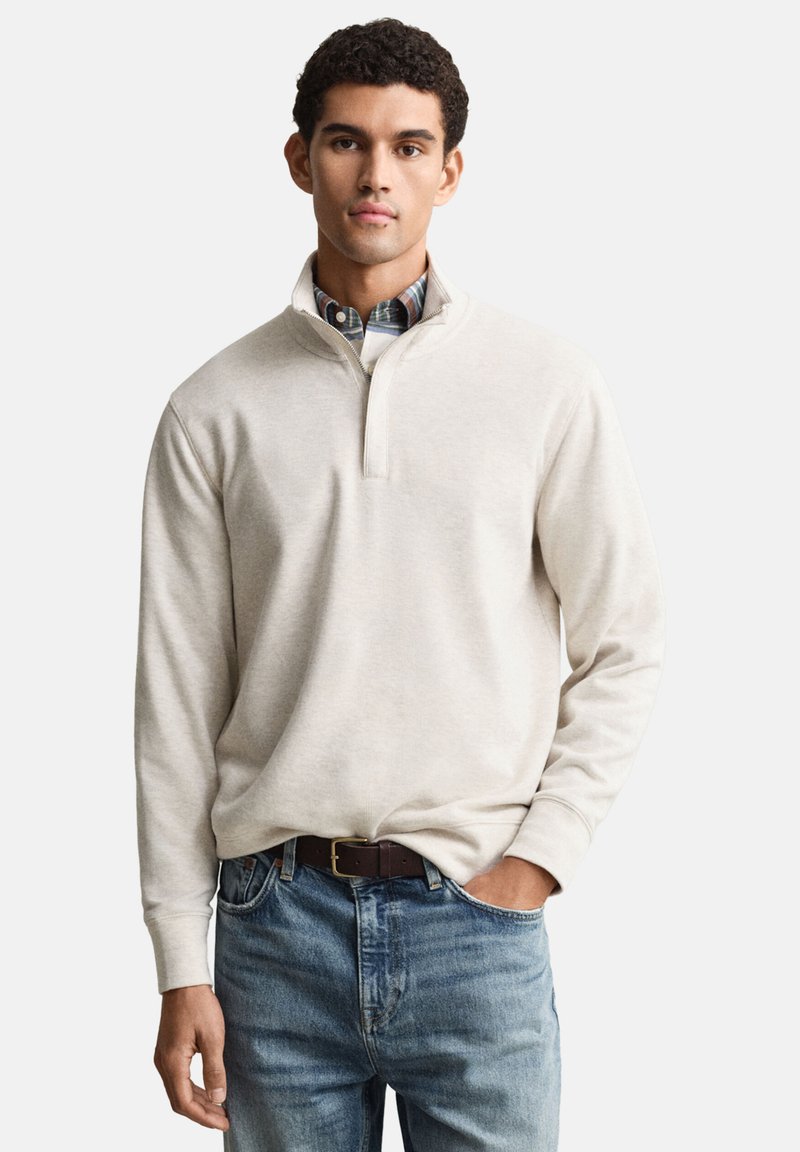 Maglione beige chiaro con collo a zip corta, realizzato in tessuto morbido. Indossato sopra una camicia a quadri, abbinato a jeans di denim blu e cintura marrone.