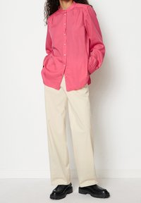 Camicia fucsia con bottonatura, colletto alto, maniche lunghe e dettagli arricciati. Abbinata a pantaloni larghi color crema e scarpe nere.