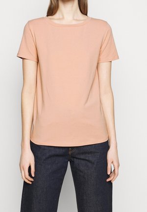Femme portant un t-shirt rose clair uni à manches courtes et un jean en denim bleu foncé, debout devant un fond blanc.