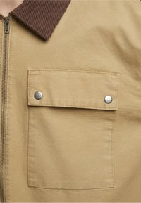 Khaki jakk, millel on pruun kallutatud kaelus, rinnakott hõbedaste näpu nupudega ja sile kangas. Zipper sulgemine.