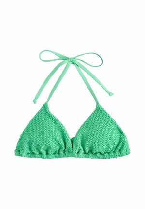 Woman in green bikini and sunglasses standing on rocky shore holding pink striped beach bag near ocean.