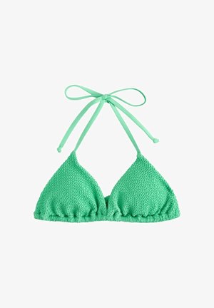 Femme en bikini vert et lunettes de soleil debout sur une rive rocheuse tenant un sac de plage rose rayé près de l'océan.