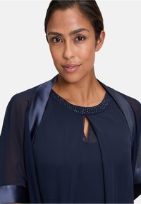 Navy blouse with sheer sleeves and a round neckline. Features a keyhole opening and decorated neckline with small, embedded rhinestones.