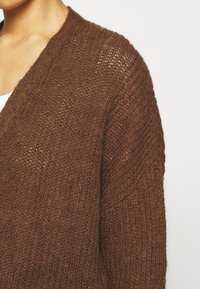 Hnědý pletený cardigan s volnou tkaninou, designem ve tvaru V a mírně oversized střihem. Textura je jemná a vzdušná s výrazným prošíváním.