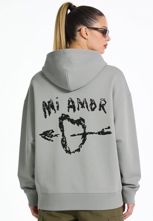 Woman wearing gray hoodie with black text "Mi Amor" and an arrow through a heart graphic on the back, standing in profile with sunglasses.
