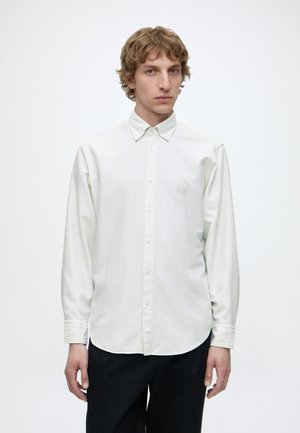 Jeune homme aux cheveux blonds bouclés portant une chemise rayée claire boutonnée et un pantalon noir sur un fond blanc uni.