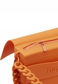 Borsa arancione in gomma con superficie testurizzata, dotata di una tasca con cerniera e una tracolla a catena robusta. Forma quadrata con bordi arrotondati.