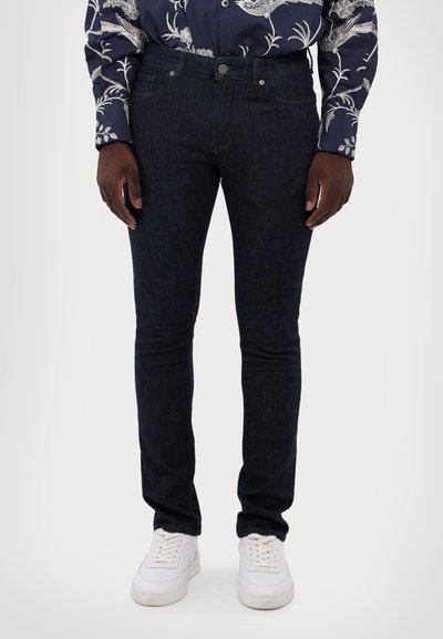 Jeans de mezclilla azul oscuro, con un corte slim, diseño de cinco bolsillos y detalles de costura sutiles. Combinados con zapatillas blancas.