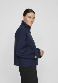 Marineblå ulljakke med høy krage, gullknapper og sidelommer, som har en firkantet silhuett og strukturert skulderdesign.