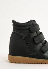 Tenisi negri cu toc wedge, cu trei benzi Velcro, guler căptușit și talpă din cauciuc de tip gumă, pe un fundal deschis.