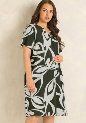 Woman in a knee-length, black and white leaf-patterned dress with short sleeves, standing against a plain light background.