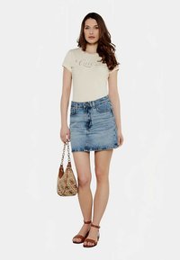 T-shirt beige clair avec logo "Guess", mini-jupe en denim bleu, sandales marron à brides et sac à main beige à motifs avec bandoulière chaîne.