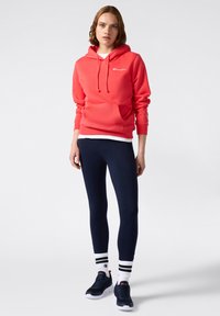 Sudadera de coral con bolsillo frontal y cordones, combinada con leggings navy y zapatillas negras. El modelo lleva calcetines a rayas y está de pie frente a un fondo liso.