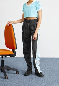 En person som bär svarta och vita Adidas träningsbyxor och en ljusblå croppad Adidas-tröja, står bredvid en orange kontorsstol.