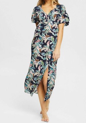 Vrouw draagt een donkerblauwe bloemenprint wikkeljurk met pofmouwen en een voorzijde splitting, staat blootsvoets tegen een effen achtergrond.