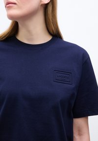 Γυναίκα που φοράει απλό μπλε σκούρο T-shirt Lacoste με στρογγυλή λαιμόκοψη και μικρό ορθογώνιο ανάγλυφο λογότυπο στο στήθος.