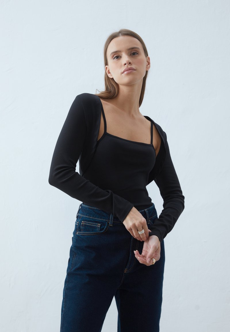 Svart långärmad topp med fyrkantig halsringning som bärs över en åtsittande linne, tillsammans med mörkblå denimjeans. Slät textur, avslappnad passform.