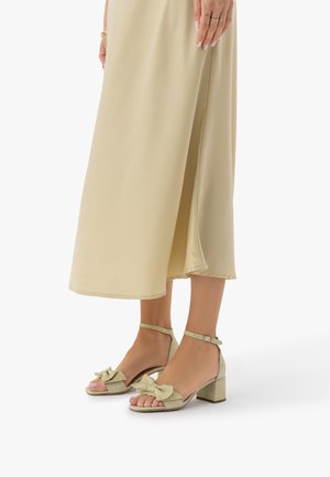 Dolne części nóg w bladożółtej spódnicy midi oraz dopasowanych sandałach na słupku z paskami wokół kostek i kokardkami na czubkach palców.