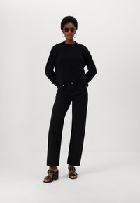Maglione corto a costine nero, jeans neri a vita alta e sandali neri con lacci e dettagli dorati. La modella posa su uno sfondo bianco.