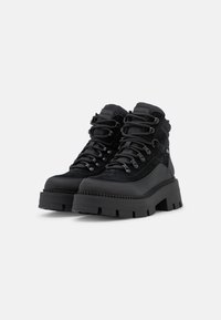 Czarne wysokie buty trekkingowe z grubymi podeszwami o dużych wypustkach, z przodem sznurowanym, metalowymi oczkami i wyściółką na kostkach na białym tle.