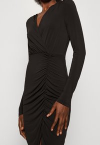 Czarna sukienka z długim rękawem i głębokim dekoltem w kształcie litery V, z zebranym materiałem w talii i bocznym rozcięciem, wykonana z elastycznej tkaniny.