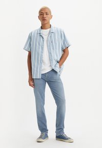 Kortermet skjorte med knapper i blå og hvite vertikale striper, sammen med lyseblå jeans og blå sneakers, stående mot en hvit bakgrunn.
