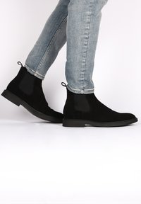 Bottes Chelsea en daim noir avec des panneaux élastiques sur les côtés, une languette de traction et une semelle en caoutchouc plate, associées à un jean skinny bleu clair.