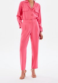 Ensemble deux pièces rose vif comprenant une chemise à manches longues avec poches boutonnées et un pantalon ajusté à plis. Tissu lisse et coupe slim.